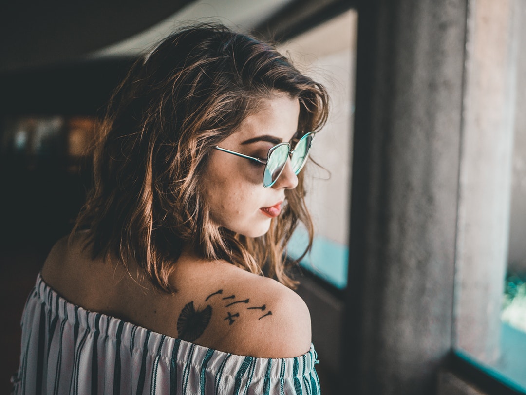 A woman in sunglasses wears an off-the-shoulder top, which shows a tattoo on her back.