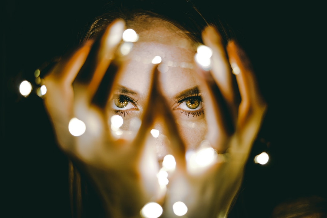 A close-up of woman's eyes through her hands surrounded by fairy lights.