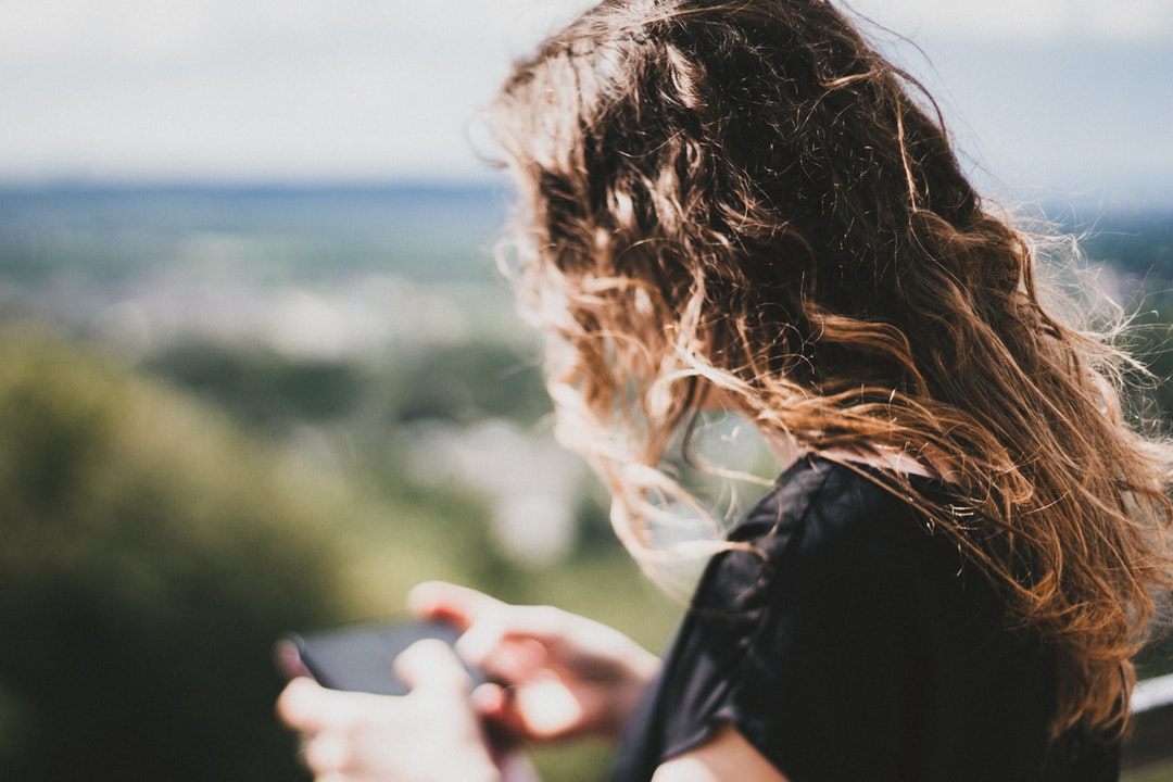 A woman with brown curly hair looks down to use her cell phone outdoors