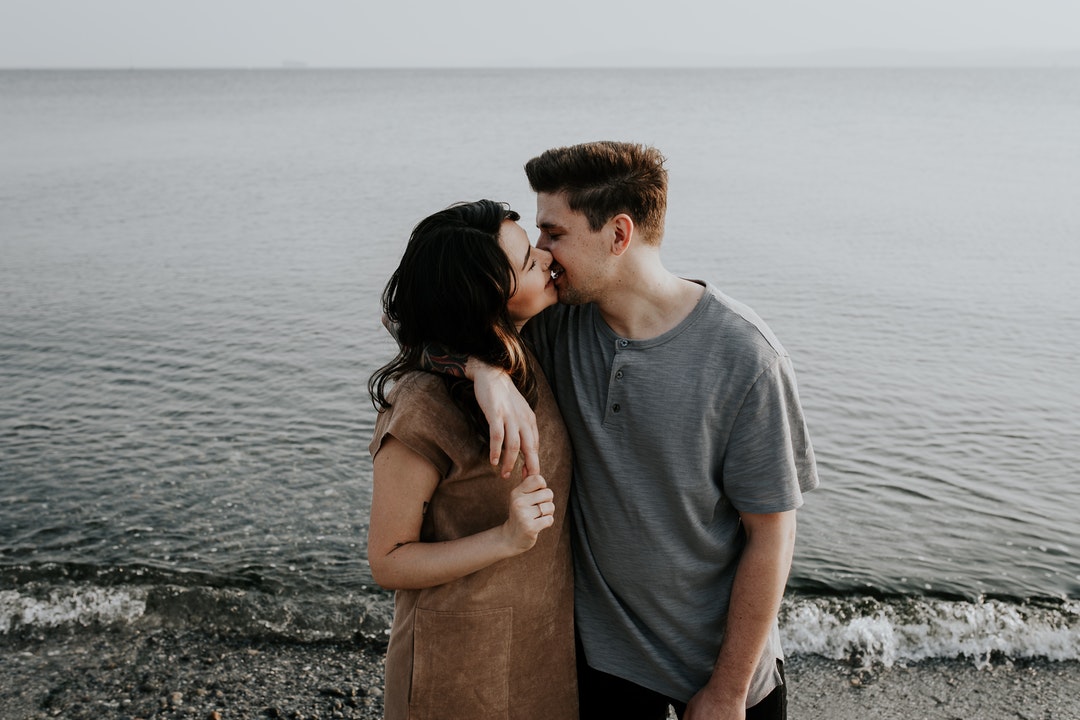 A man with his arm wrapped around a woman, reaching in passionately for a kiss while standing near the water on a beach.