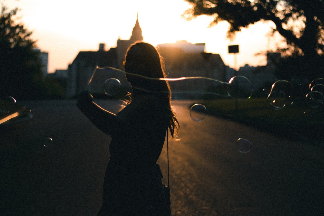 silhouette photo of woman holding bubbles near building during golden hour