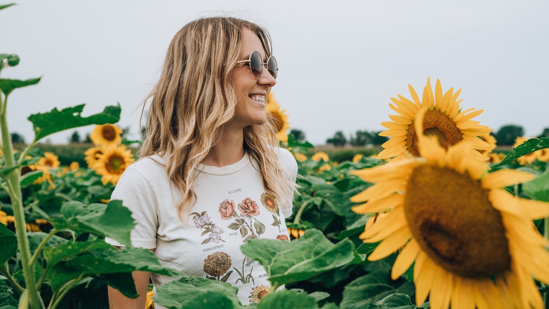 woman in white and multicolored floral shirt standing and smiling on yellow sunflower field
