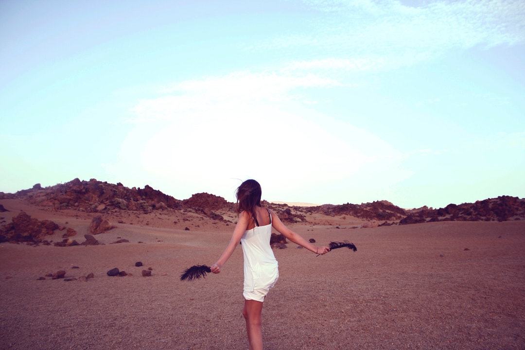 A carefree young woman with a black feather in either hand in the middle of a barren rocky plain