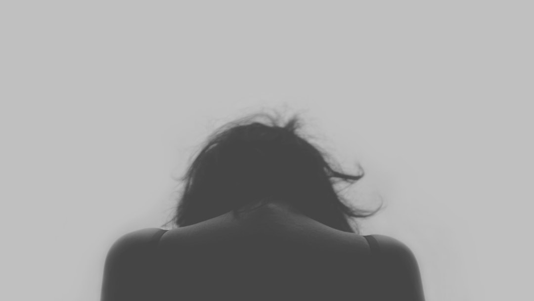 A black and white rear shot of the back of a woman's head and back, with her head bowed down in sadness