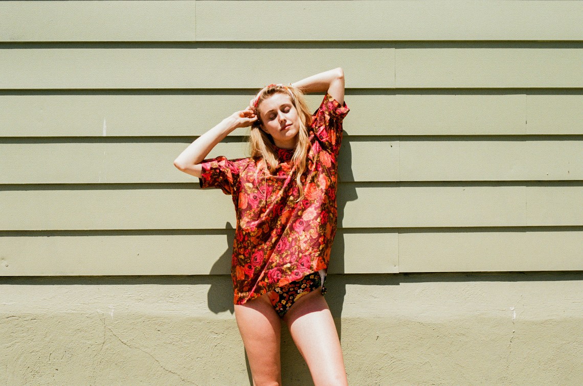 Woman in an orange shirt and leather purse stretches out in front of a wall, looking carefree and young