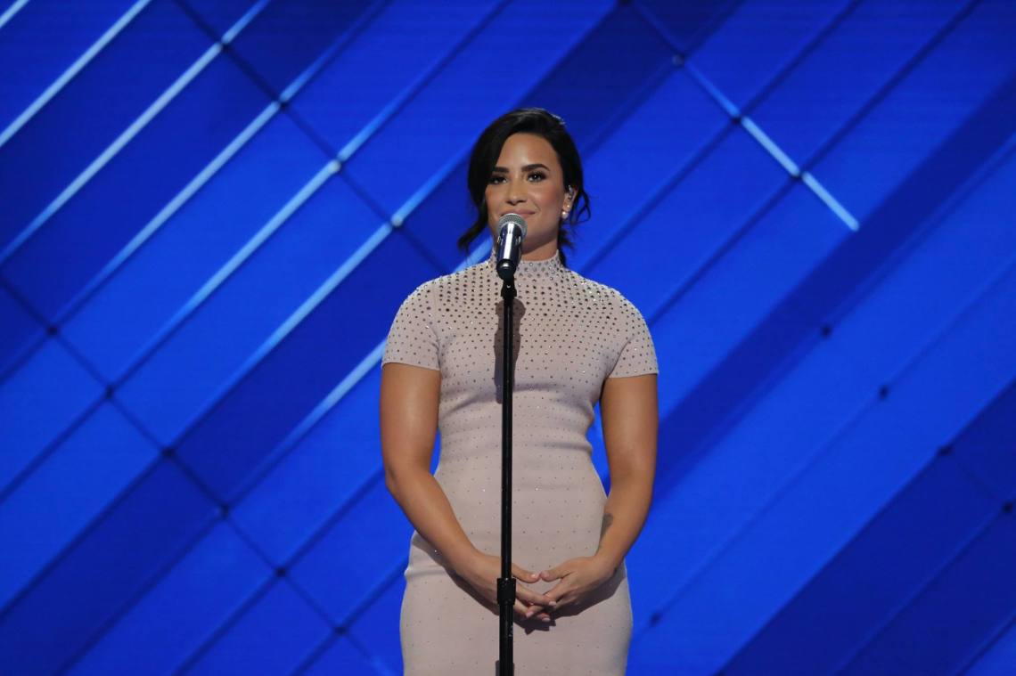 Demi Lovato stands on stage where a white dress in front of a blue background for some event