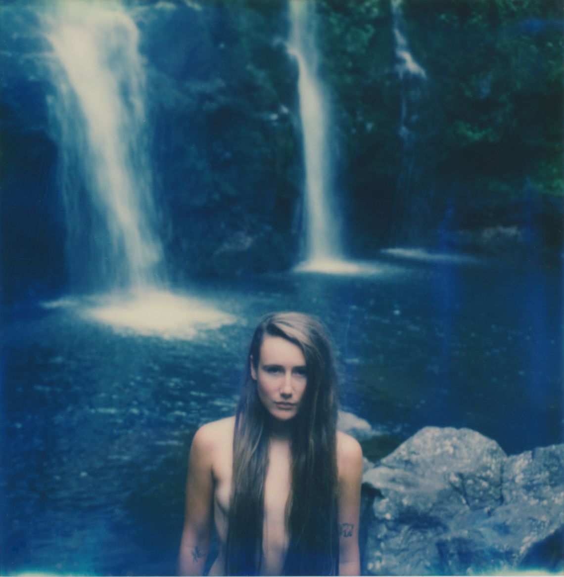 Blue tint on nude girl standing by waterfalls with long brown hair