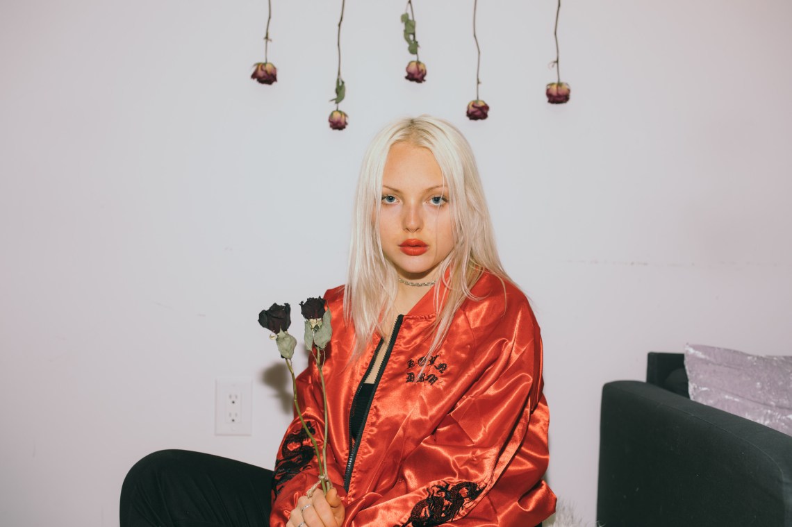 Woman sits in a room underneath hanging roses, staring at the camera