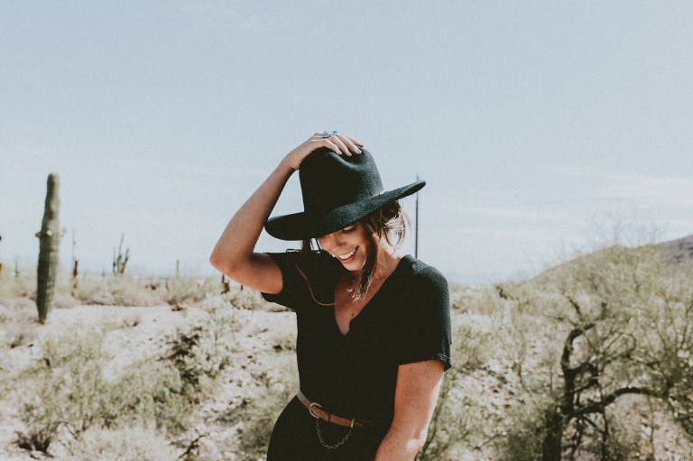 Girl in all black in the desert holding down the hat on her head, cactus cacti