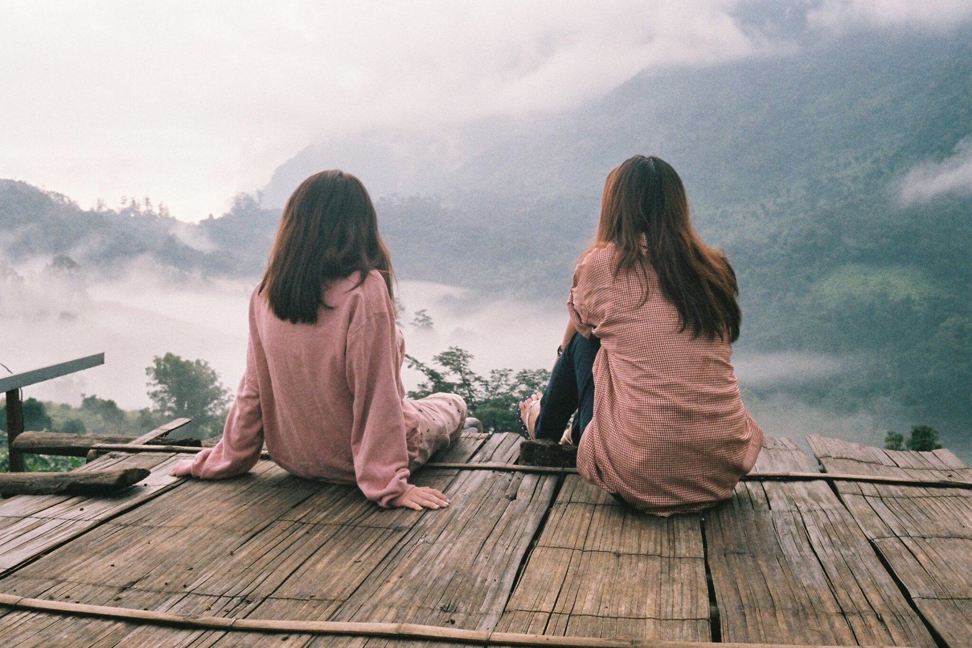 Two brunette girls in matching dusty rose pink shirts sitting on a rickety wooden platform overlooking misty mountains