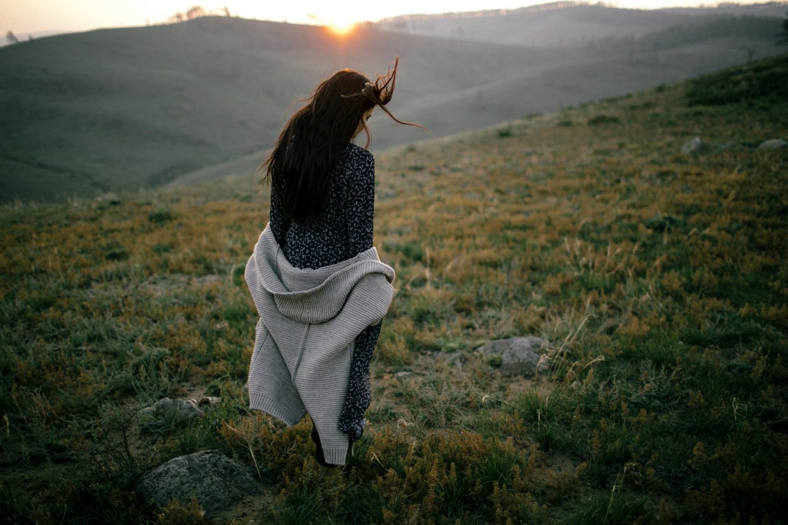 Girl taking a leisurely walk through rocky rough grasses and the sun is setting over the hills