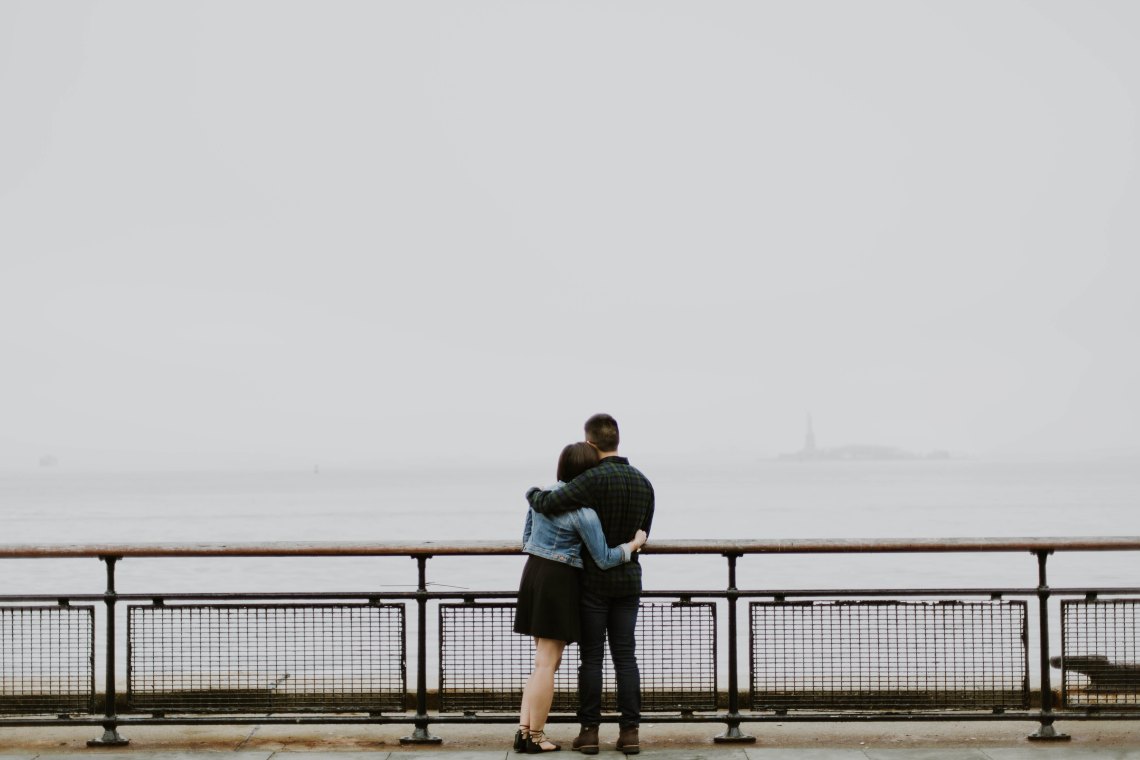 Woman and man hold each other while they stand on a bridge, staring out into the unknown together