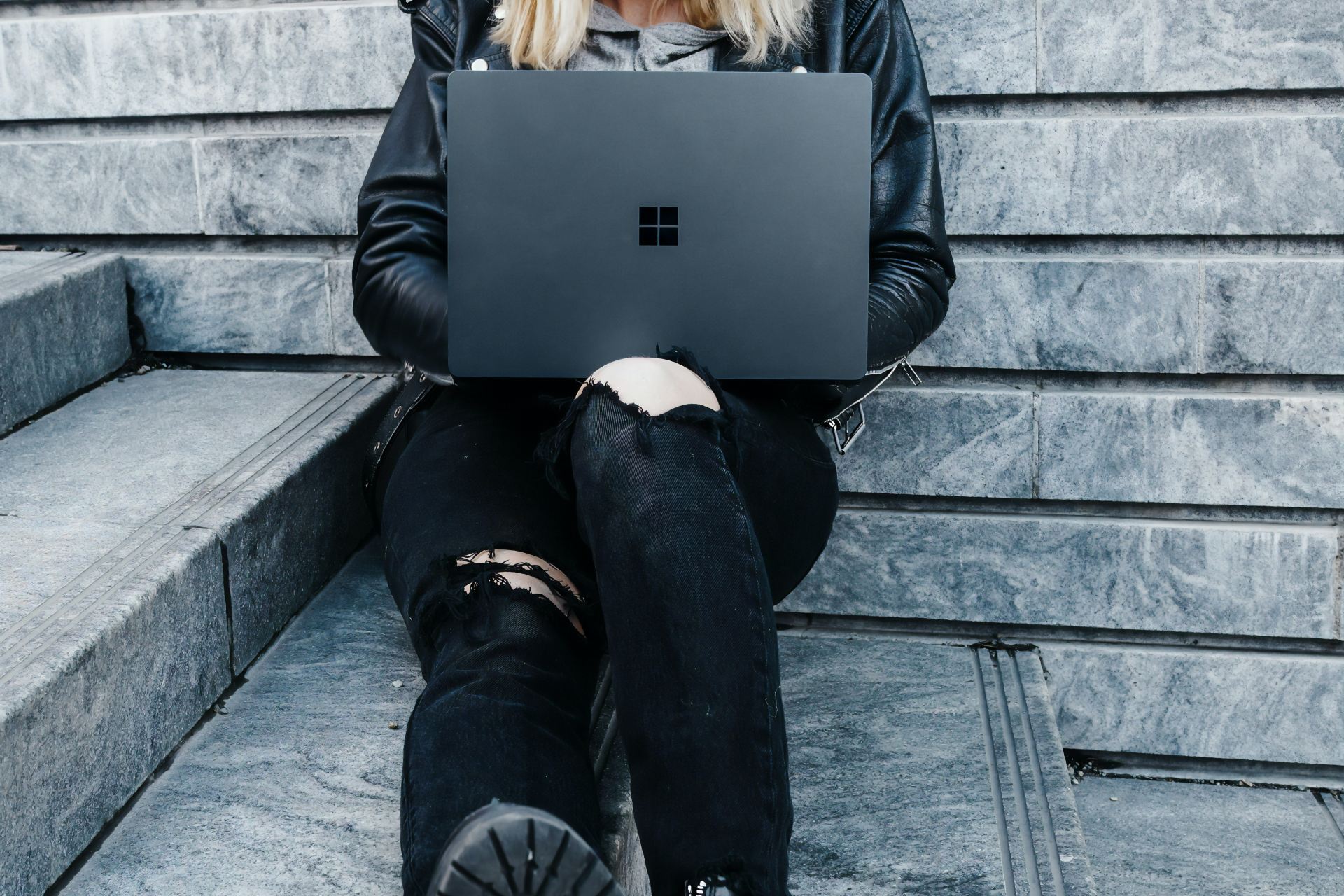 woman in black jacket and black pants sitting on concrete stairs usingMicrosoft Surface Laptop