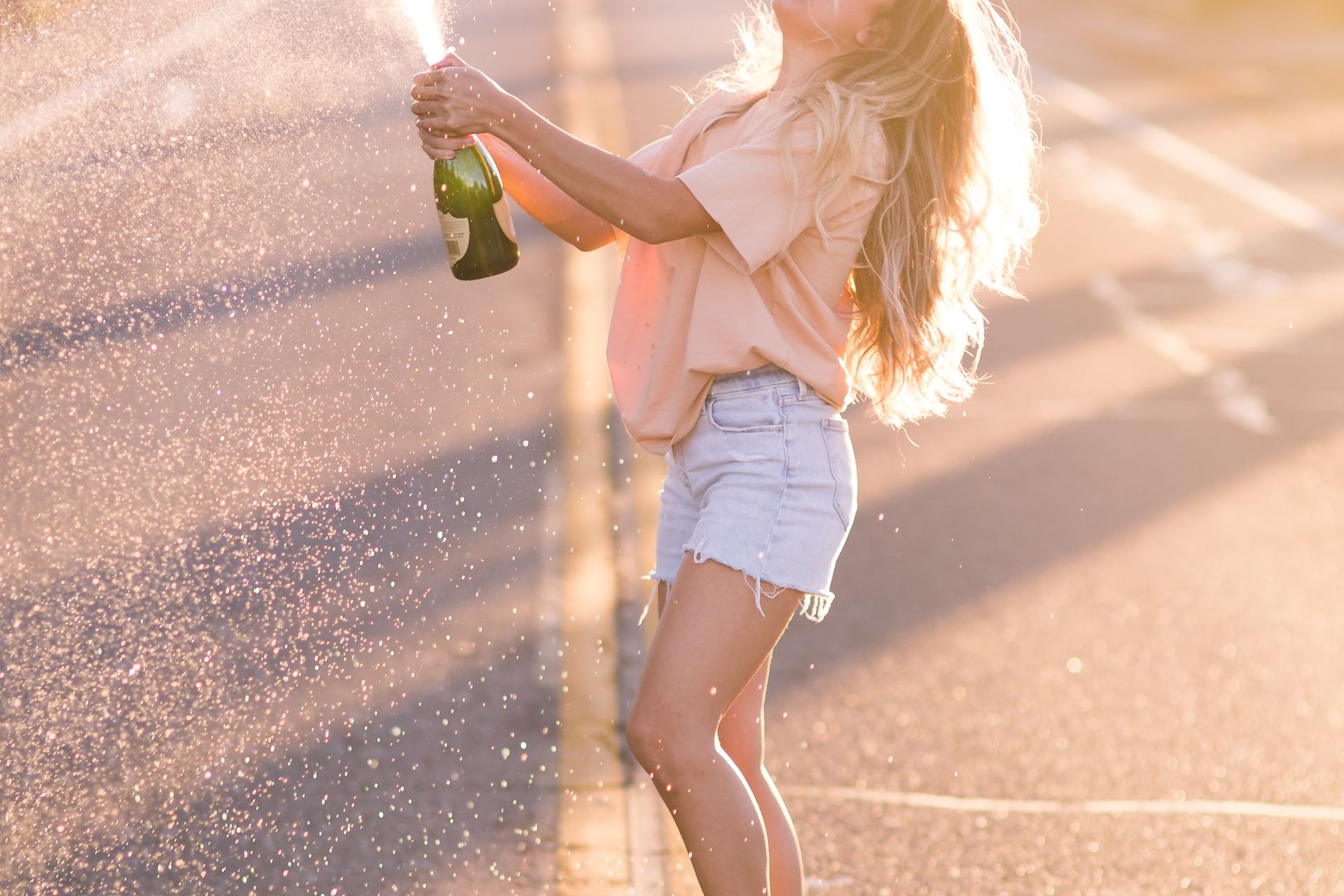 woman in white shirt and blue denim shorts holding green bottle standing on road during daytime