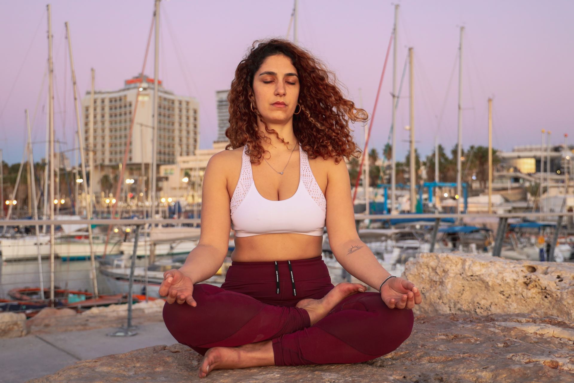 woman wearing white sports bra and red leggings sitting while doing yoga viewing buildings and boats on body of water during daytime