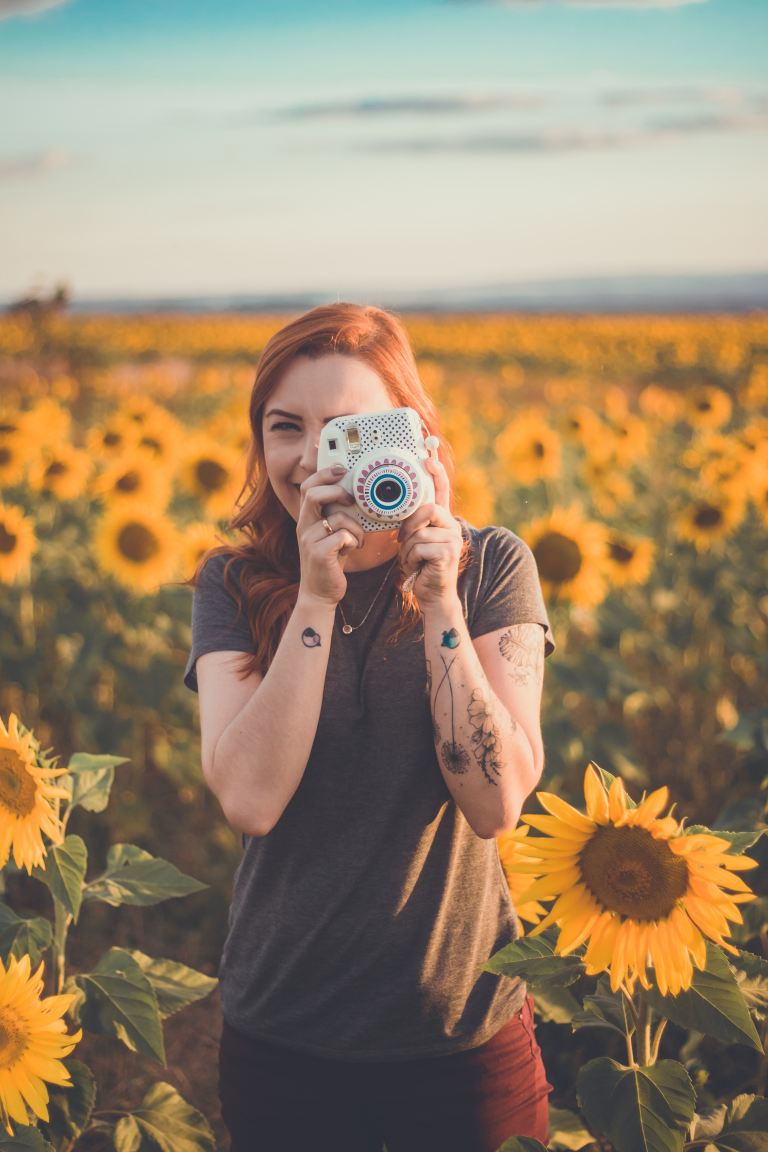 woman holding white instant camera standing on meadow of Sunflowers