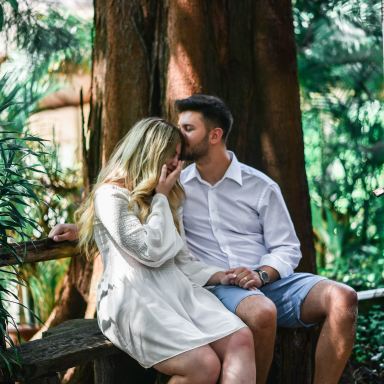 man in white dress shirt kissing a woman in white long-sleeved dress while sitting on bench beside brown large tree at daytime