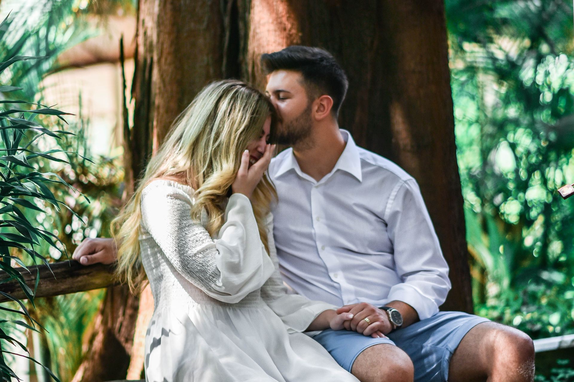 man in white dress shirt kissing a woman in white long-sleeved dress while sitting on bench beside brown large tree at daytime